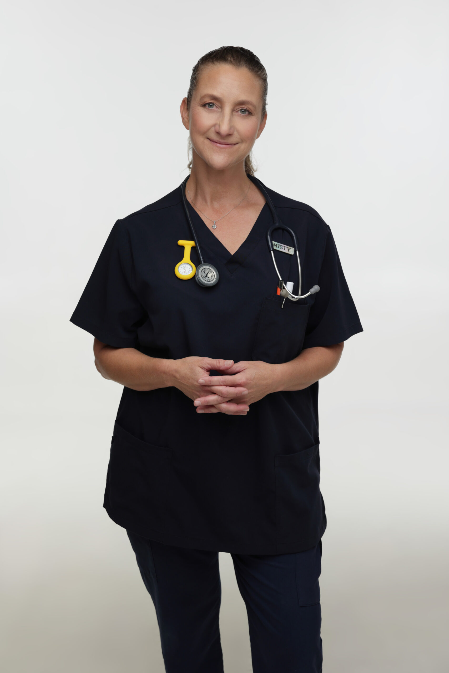 A woman wearing dark medical scrubs stands against a plain background, smiling gently. She has a stethoscope round her neck and a yellow nurse's watch clipped to her top. Her hands are loosely clasped in front of her.