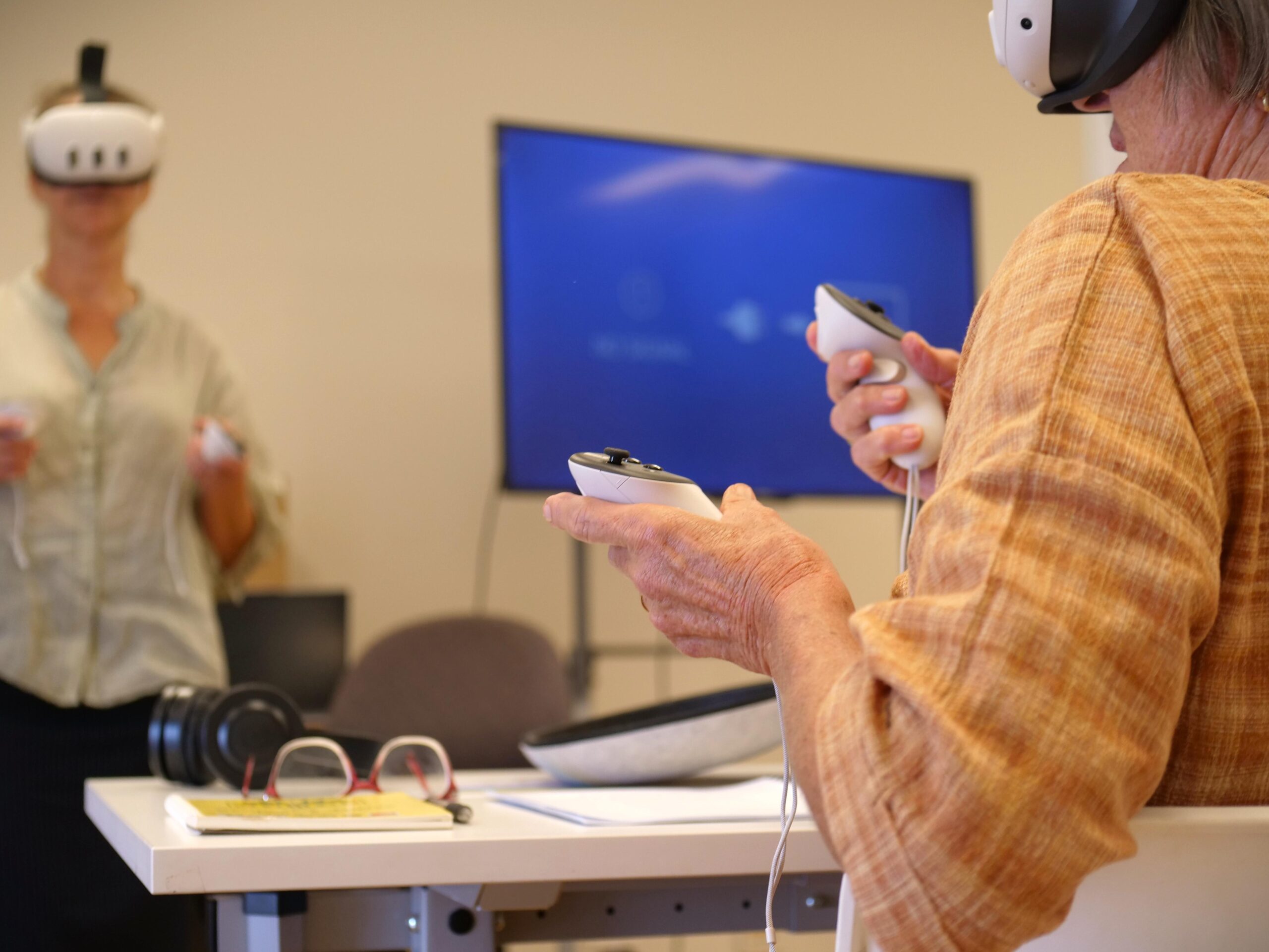 Two people use virtual reality headsets and controllers in a room with a TV screen and desk. A pair of spectacles, a notebook, and headphones are on the desk in the foreground.