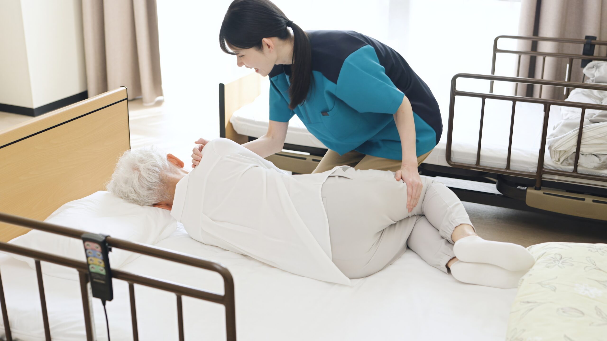 A carer assists an elderly person lying on their side in a bed, gently supporting their back and legs, in a well-lit room with another bed nearby.