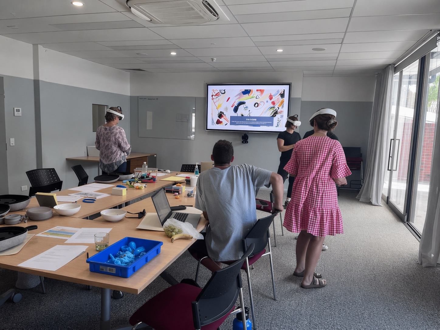 Four people in a bright meeting room stand around a table with papers, blue LEGO pieces, and other materials. A large screen displays colourful graphics. The room has glass doors and modern furnishings.