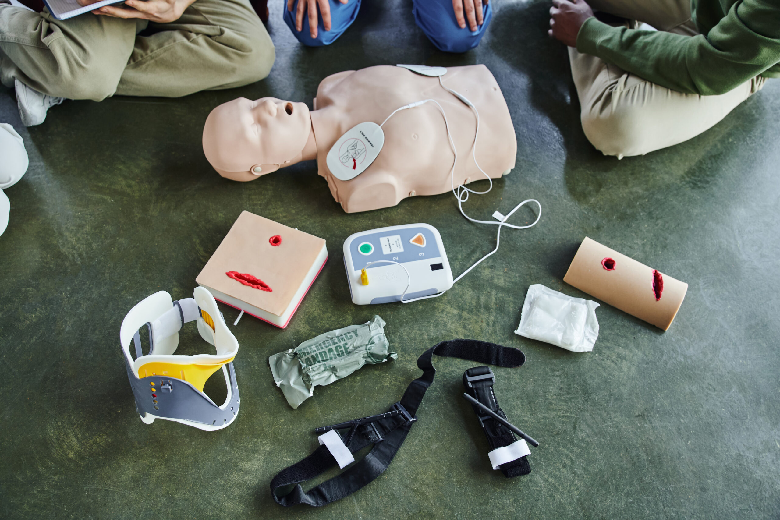 Three people sit around a medical training setup with a CPR manikin, defibrillator, wound simulation props, a neck brace, and first aid supplies arranged on the floor.