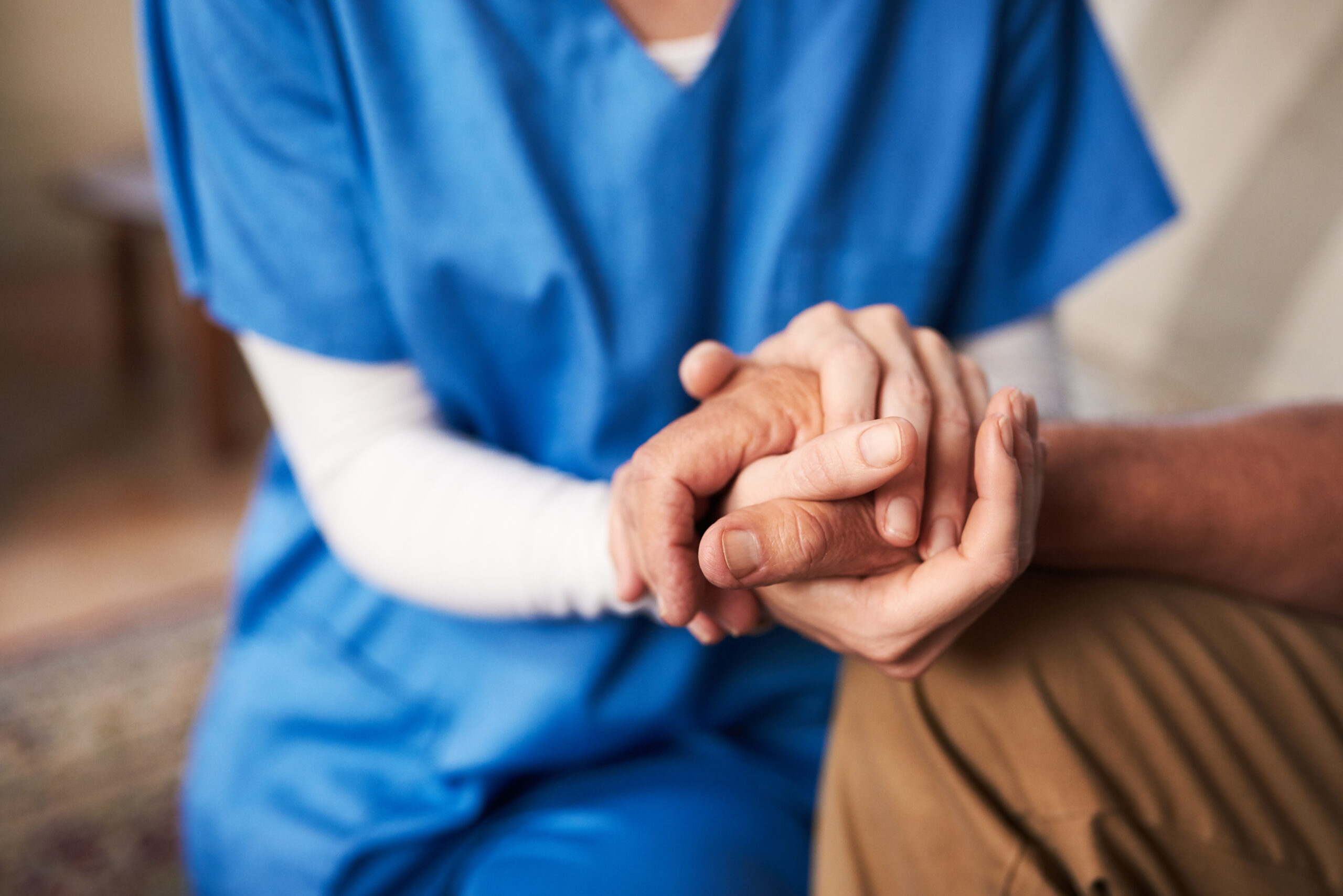 A healthcare worker in blue scrubs gently holds the hand of an elderly person, offering comfort and support.