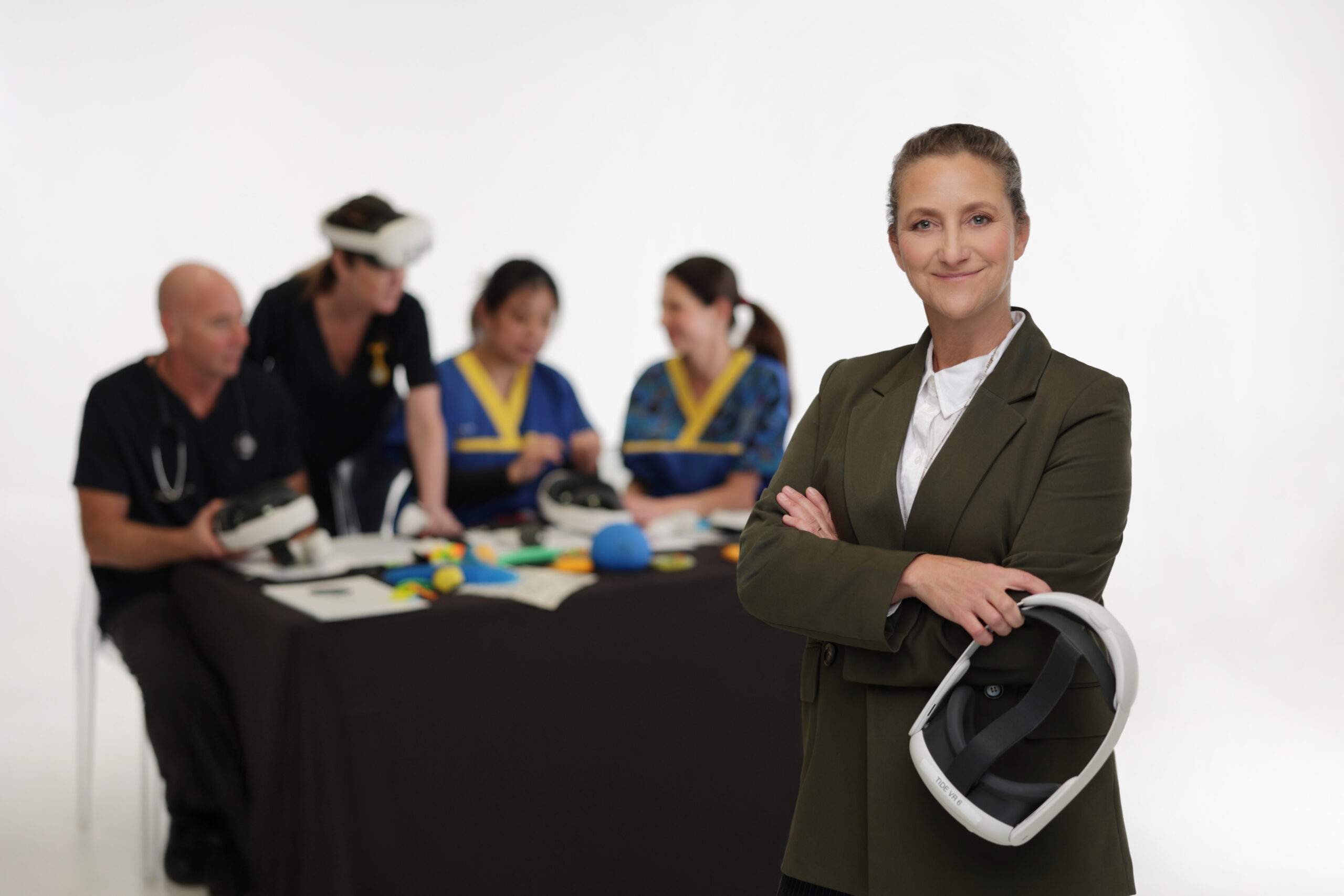A woman in a suit stands confidently holding a virtual reality headset. Behind her, four people in medical uniforms interact with VR equipment around a table. The background is plain white.